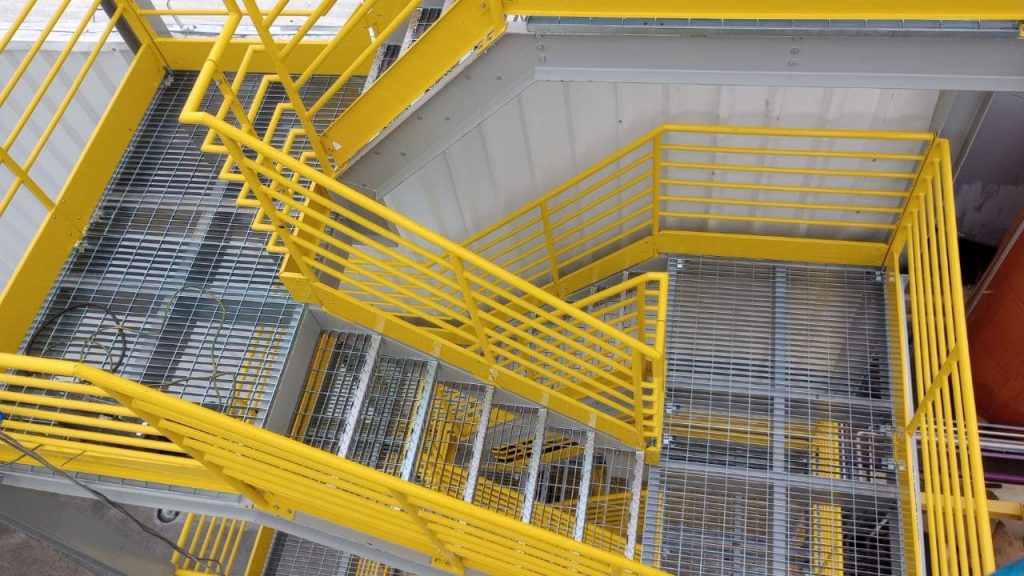 Escada metálica industrial amarela em lances com guarda-corpo e piso em grade, vista de cima